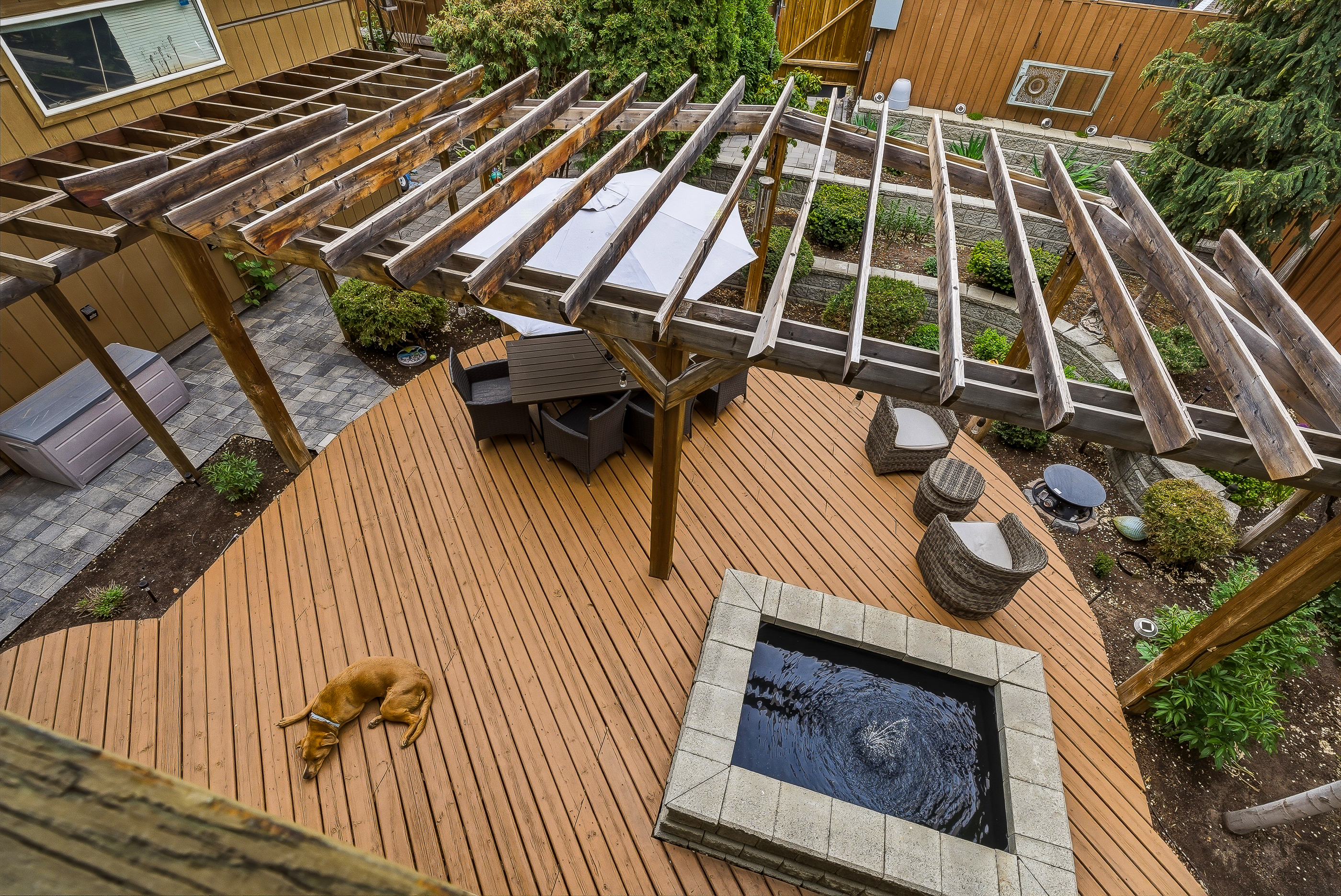 Overhead view of the back deck with pergola, water feature, raised vegetable beds, paver patio and the family dog stretched out in the sun