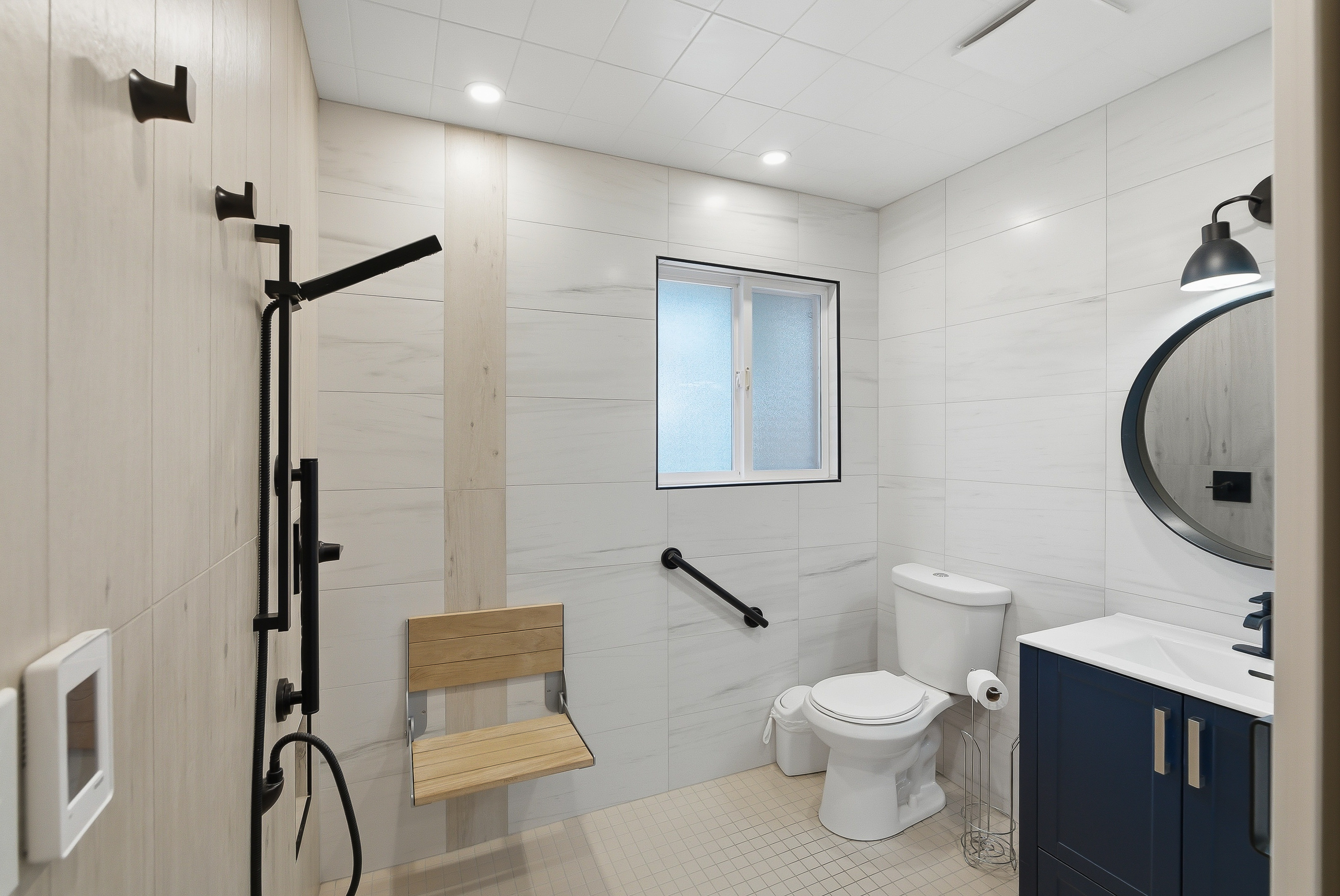 Renovated main-floor wet room at 1127 Columbia St — floor-to-ceiling tile, open walk-in shower with fold-down teak bench, grab bars and a navy vanity