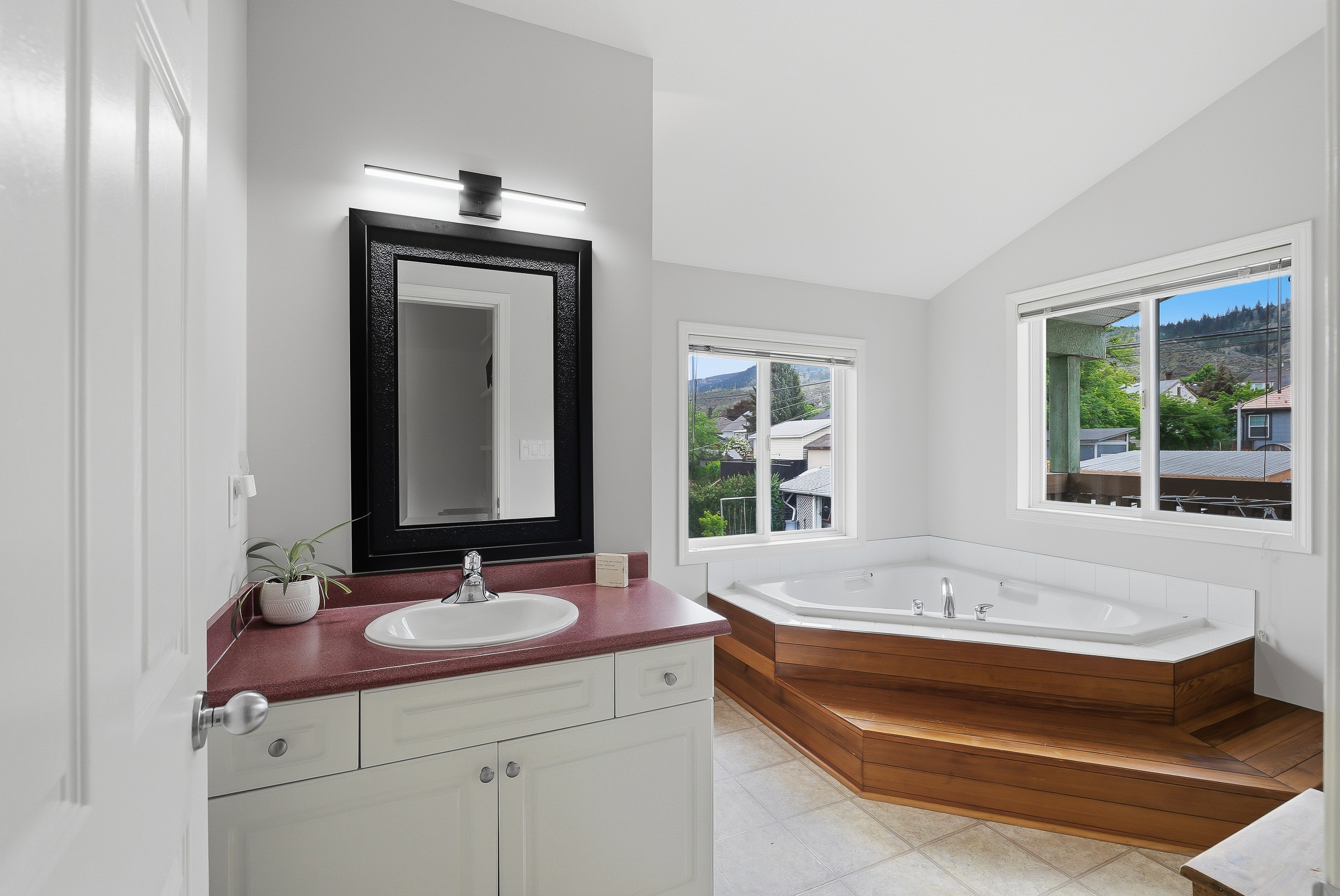 Ensuite bath with soaker tub on a teak platform, twin windows framing the Kamloops hillside, freestanding vanity and large framed mirror